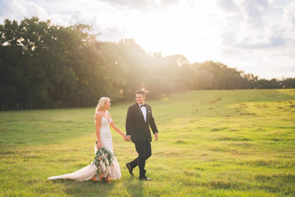 Bride and groom walking in a sunset field. They are holding hands and looking at each other smiling.