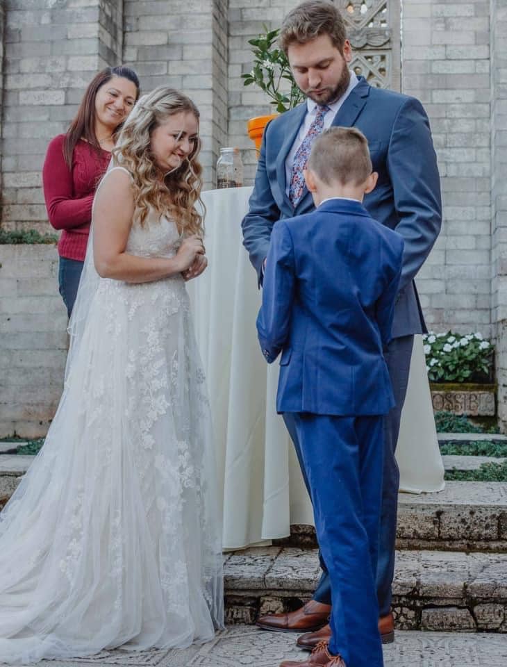 ring bearer in blue suit giving ring to groom with officiant Chrissie from Ceremonies by Chrissie looking on
