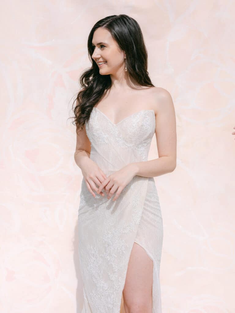 Bride wearing a slit dress with lace. She is standing in front of a pink back drop
