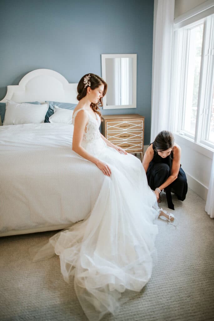 Bride sitting on a bed in her dress while a bridesmaid helps her fasten her shoes