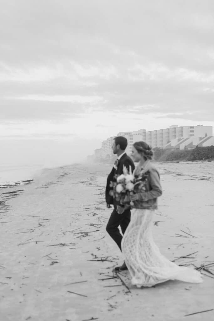 black and white photo of bride and groom on a beach looking at the waves by Joanna Moore Photography