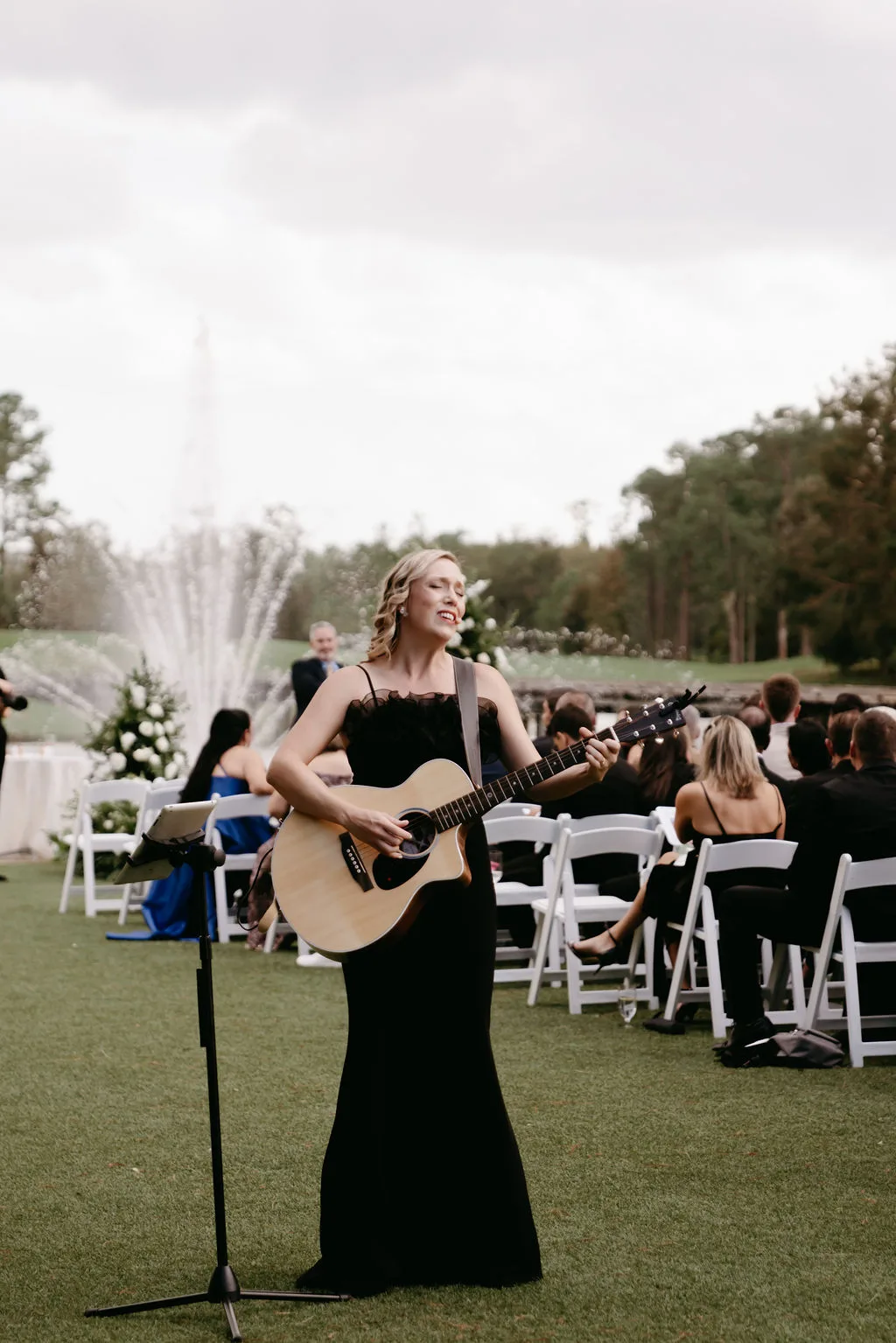 Aloha Musicians guitarist and vocalist performing in a black gown at an outdoor wedding ceremony in Orlando Florida with a fountain and white folding chairs in the background