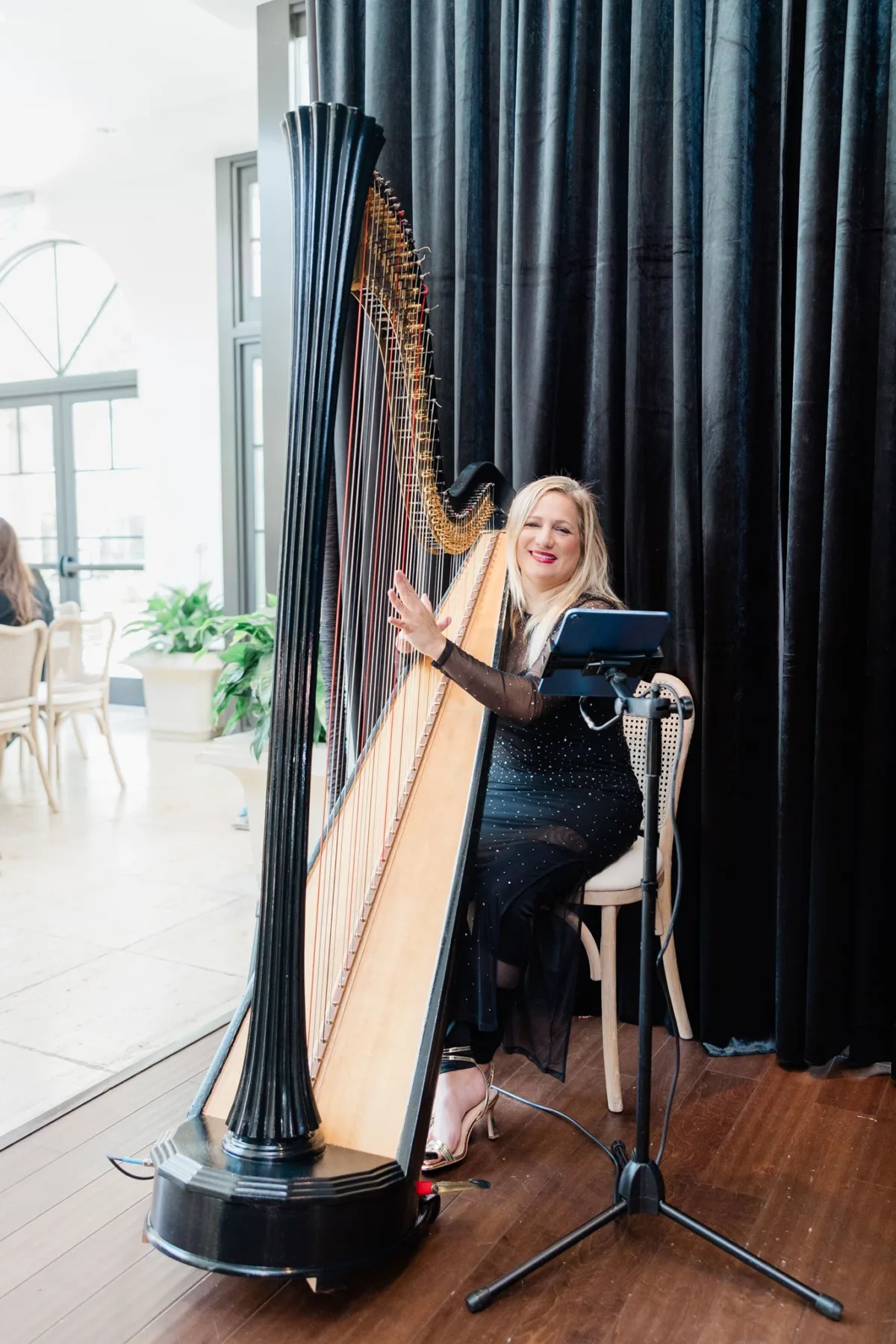 Harpist performing at an elegant Orlando wedding event, smiling beside a grand black concert harp with sheet music on a stand, provided by Aloha Musicians