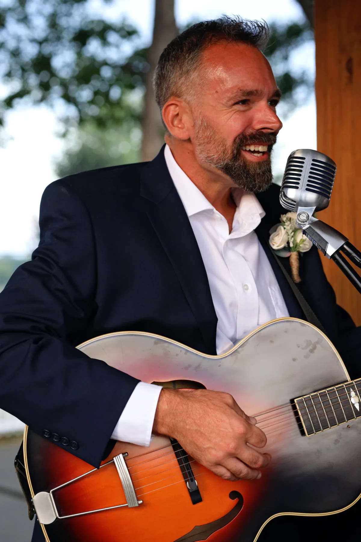 Smiling male musician playing archtop jazz guitar at a vintage-style microphone while wearing a navy blazer with boutonniere at a Central Florida wedding, performed by Aloha Musicians