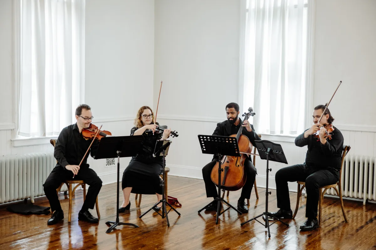String quartet performing at a bright white-walled indoor venue in Orlando Florida, featuring two violinists, one violist, and one cellist from Aloha Musicians dressed in formal black attire with music stands on a hardwood floor