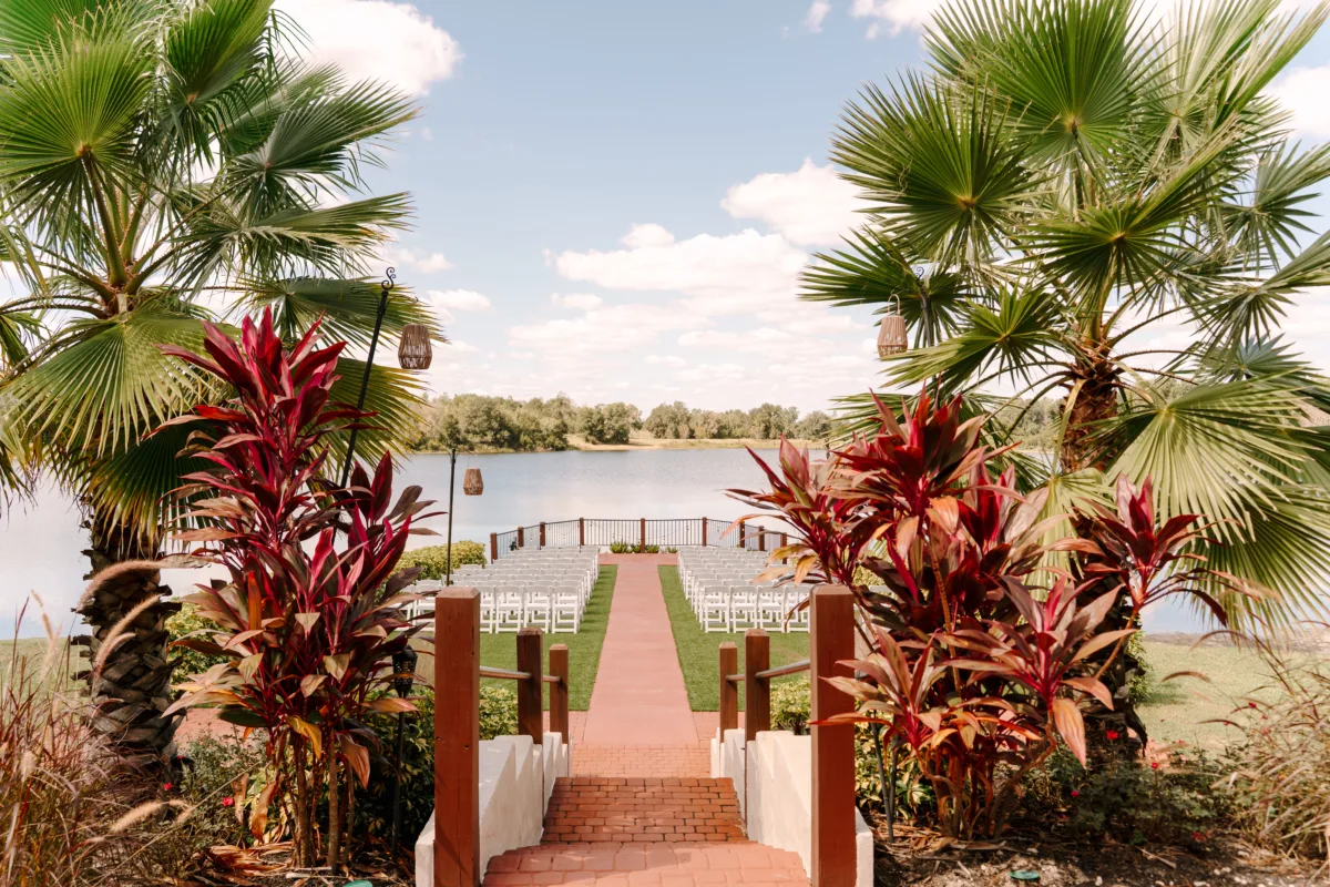 Lake Pointe ceremony site at Casani Estates featuring a brick aisle leading to white ceremony chairs, framed by lush palm trees and vibrant garden foliage, overlooking a serene lake under an open sky.