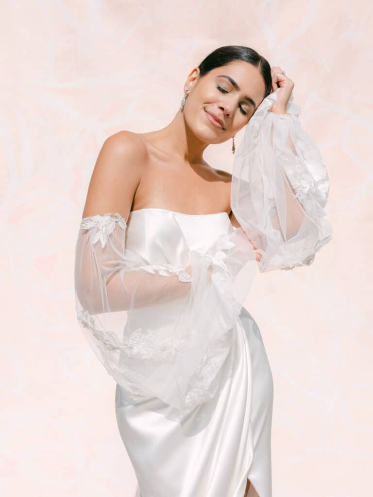 Bride in an off-the-shoulder wedding dress. Her hair is pulled back and she's against a peach color backdrop