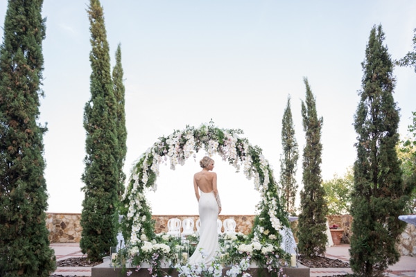 Bride posing amongst tall shrubs and in front of her wedding arch with cascading flowers