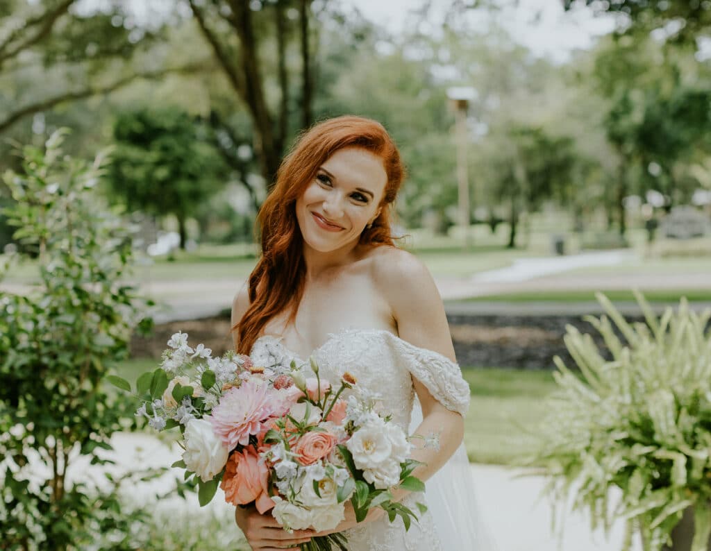 Red-headed bride among flowers. She is smiling and holding a bouquet of pink, white, and cream flowers.