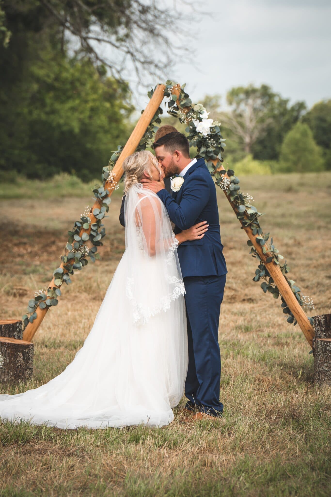 bride and groom kissing in a field under a rustic wedding altar while an officiant looks at their wedding ceremony outline