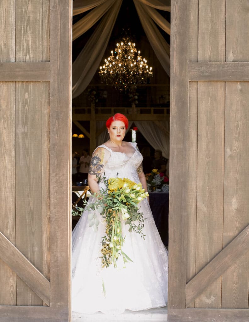 bride with bright red hair stands in open open barn doorway in wedding dress by Enchanted Bride