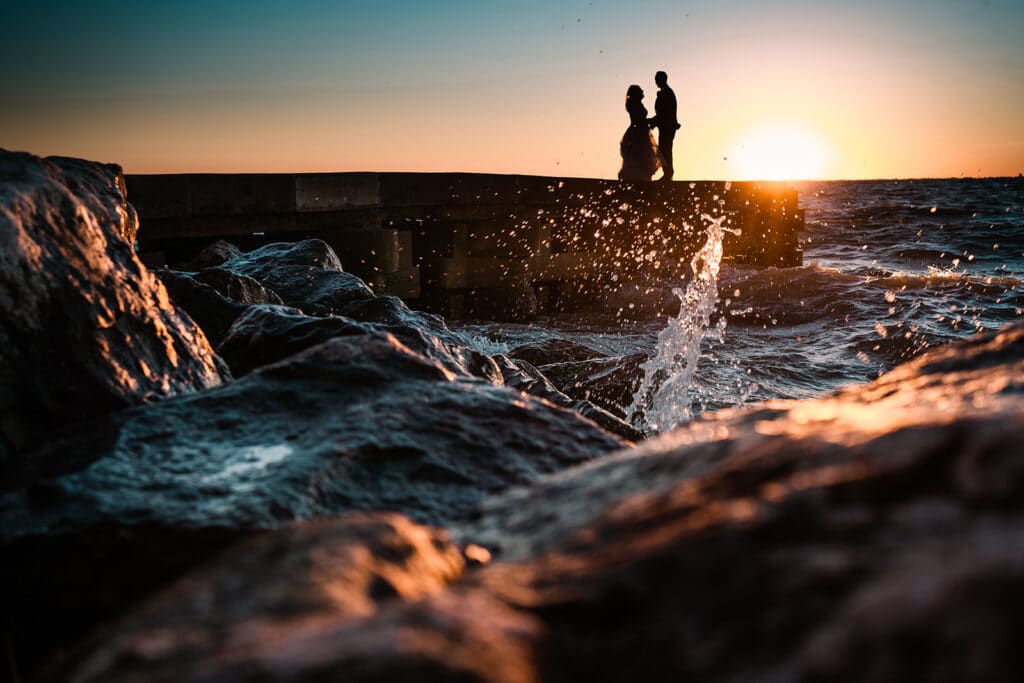 couple at the seashore with rocks and waves crashing by Roy Serafin Photo Company