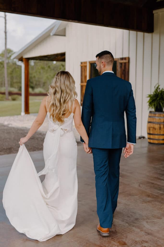 bride in slip wedding dress by Enchanted Bride and groom in navy walk toward wedding reception at a barn