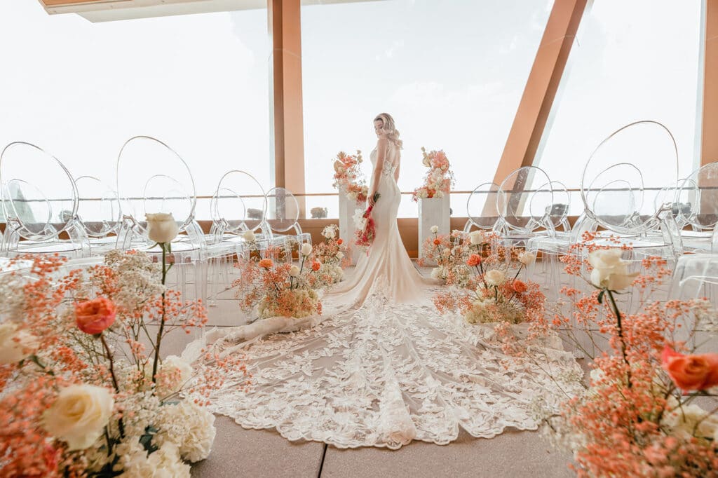 bride on balcony with lace train surrounded by white chairs by A Chair Affair Inc.and orange and white flower arrangements