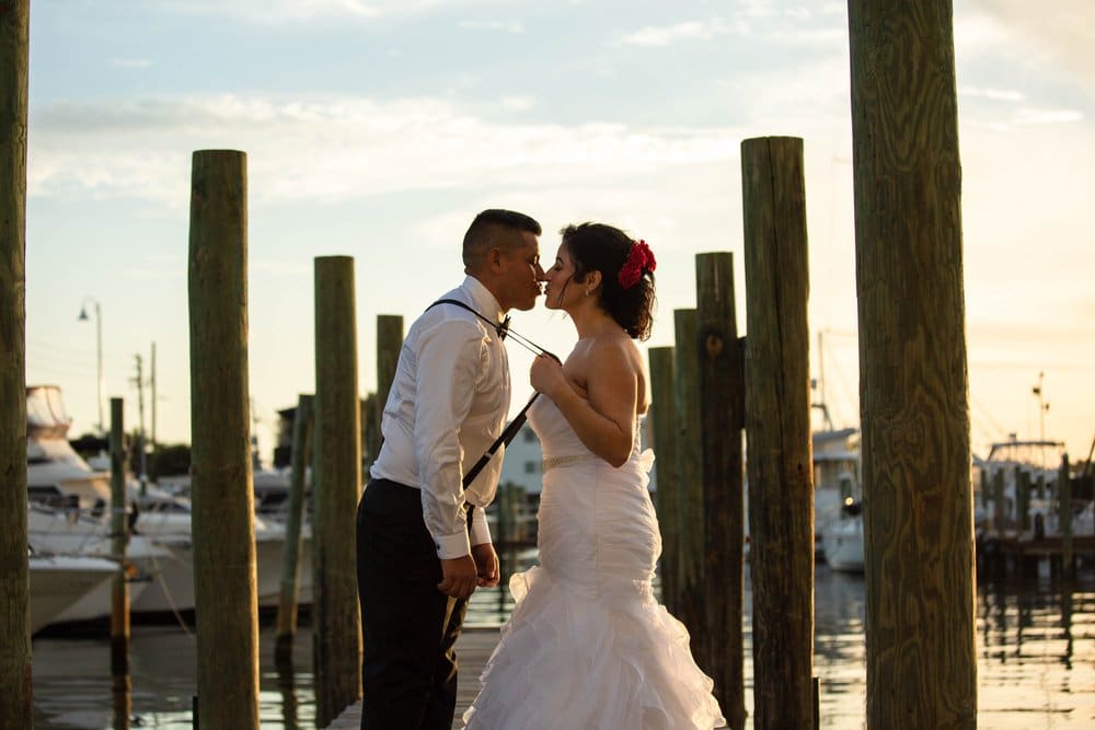 bride grasping groom by suspenders and pulling him in for a kiss on a dock in a marina