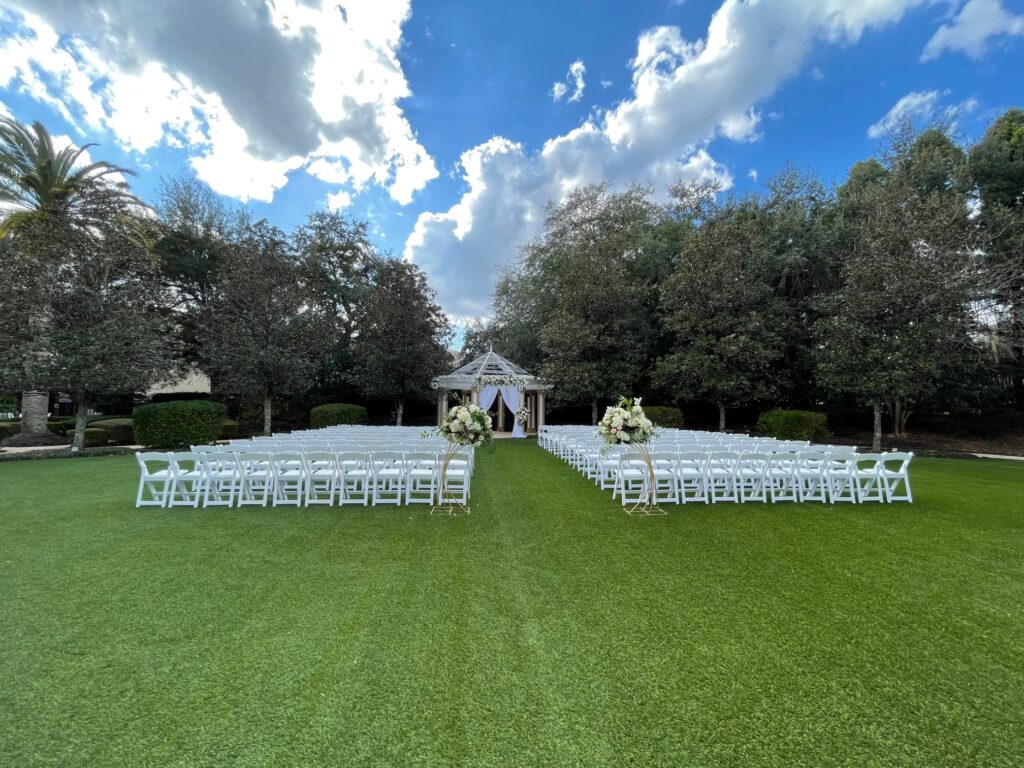 Ceremony on the lawn. There is a gazebo draped with white curtains and a few white and pink floral arrangements