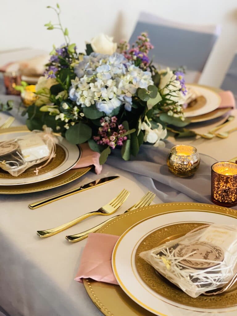 Table setting for a reception. There are flowers, gold tableware, and small gifts honoring the couple on the plates.