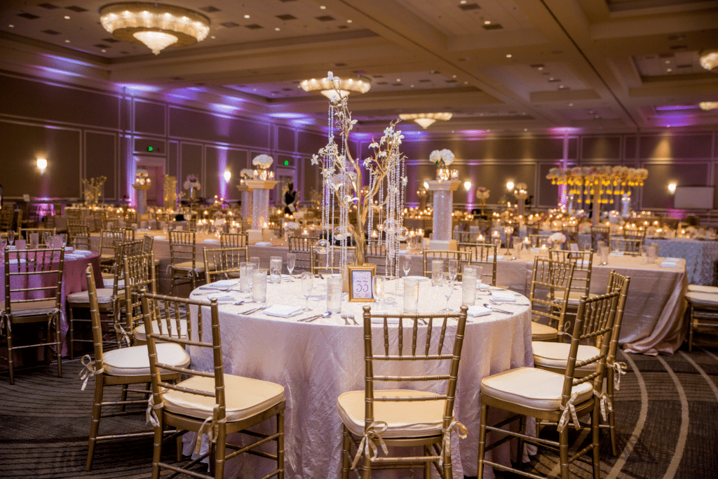 Grand ballroom set up for a wedding reception. There are crystals, large centerpieces, and gold chairs.
