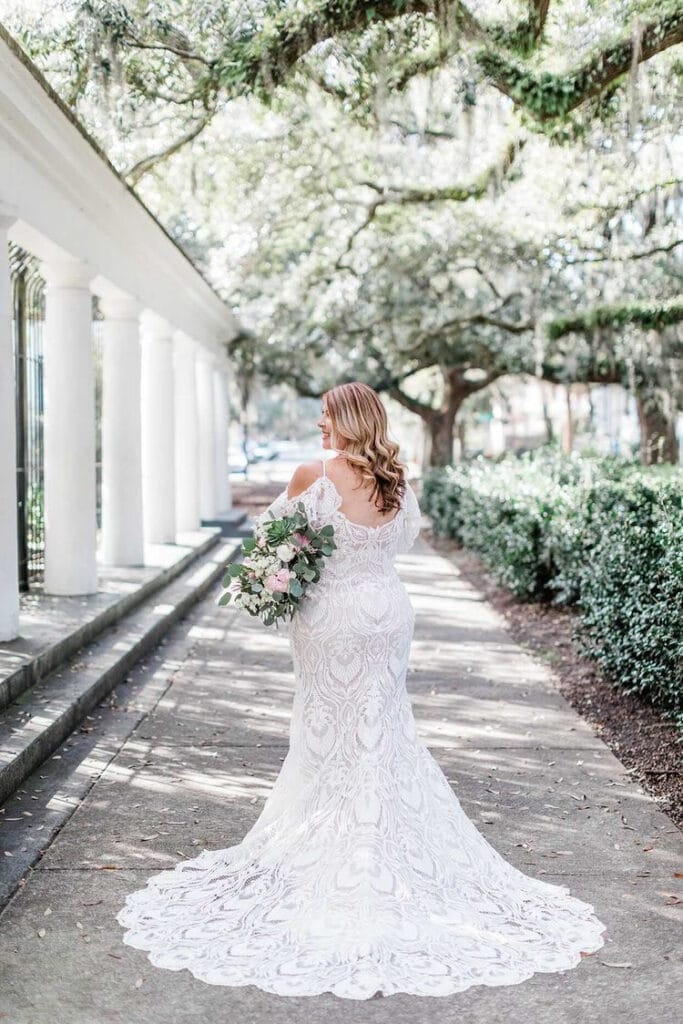 bride walking down sun dappled path with white pillars and spanish moss draped trees in wedding gown with train from the Ivy Bridal Shop