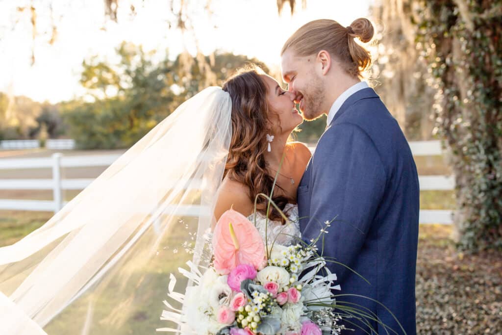 groom with man bun and bride with veil and pink and white bouquet kissing in a field surrounded by a white fence by Amy Britton Photography