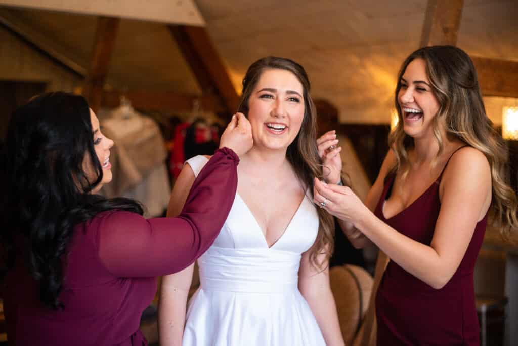 bride laughing and getting ready with her attendants in burgundy dresses, photo from Weddings By Ray