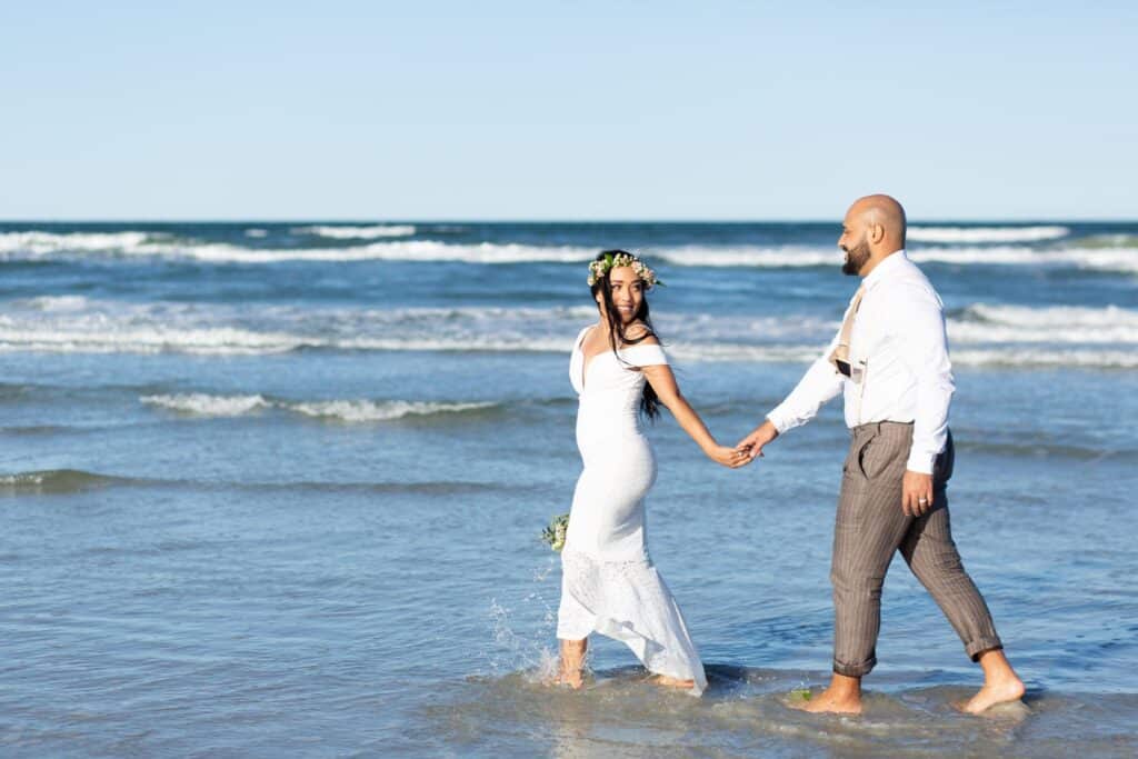 bride in wedding dress and groom in suit walking through the waves by Amy Britton Photography