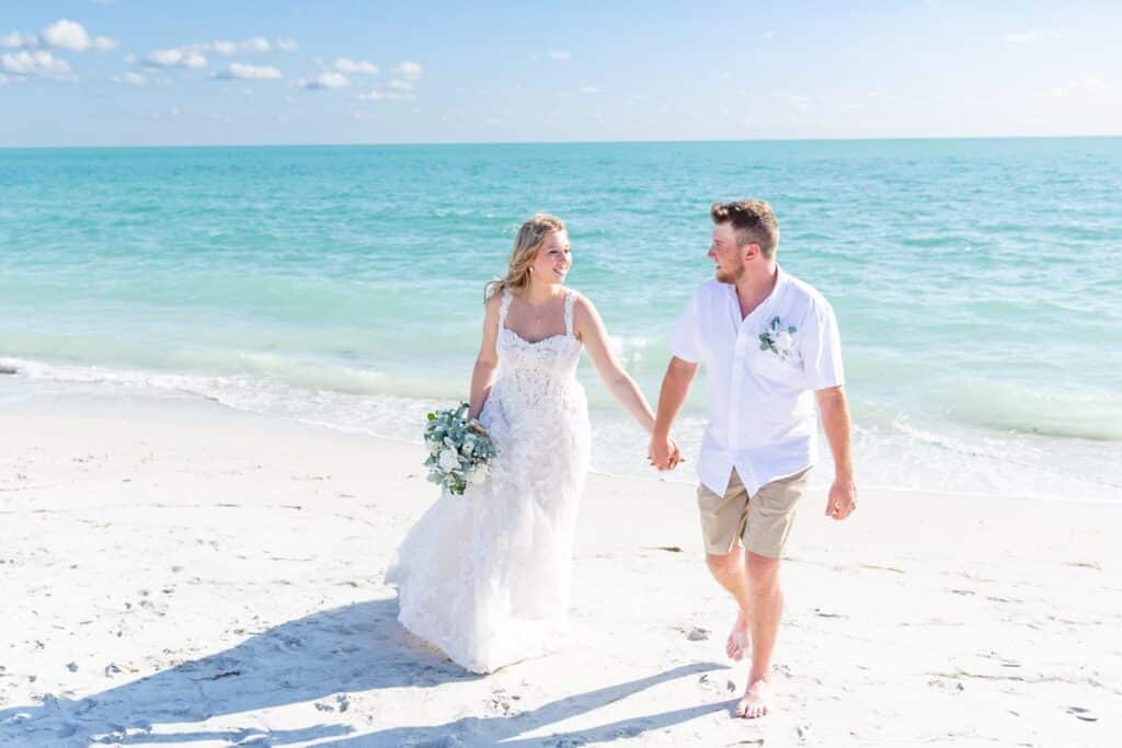 bride and groom in shorts at the beach walking in the white sand by Amy Britton Photography