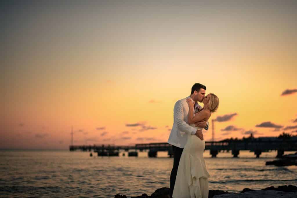 groom kissing bride at sunset on the each with pier in the background at event planned by Just Save the Date