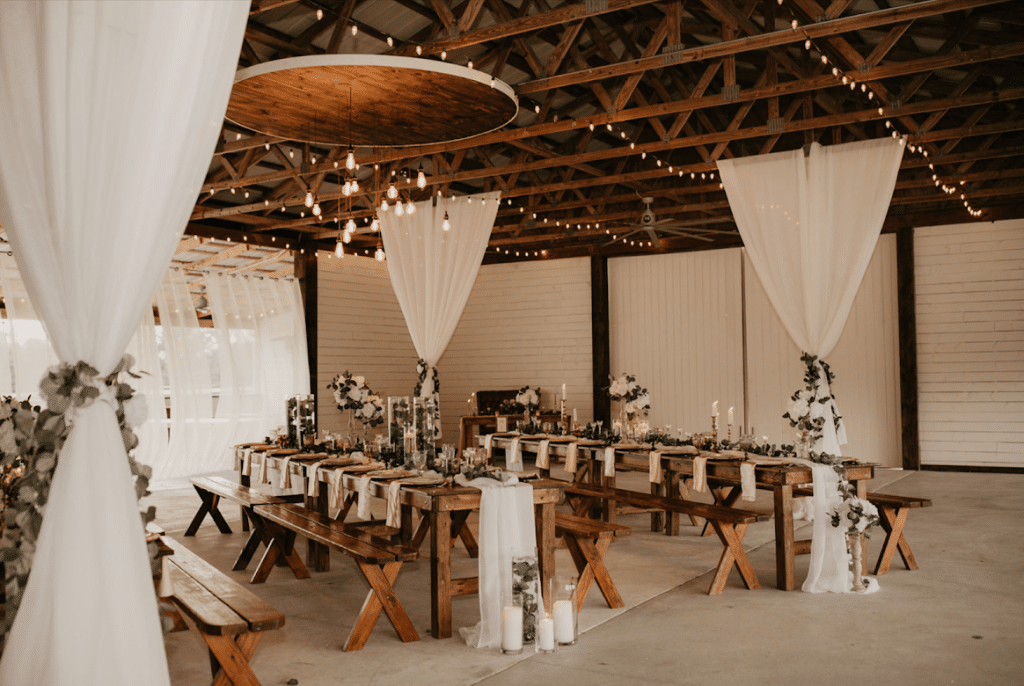 farmhouse table and benches reception area in beamed barn with white draping and chandeliers by Making It Matthews