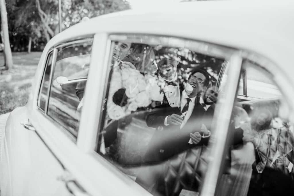 black and white photo of reflection in a car window of family waving goodbye to wedding couple leaving venue by Sydney Morman Photography