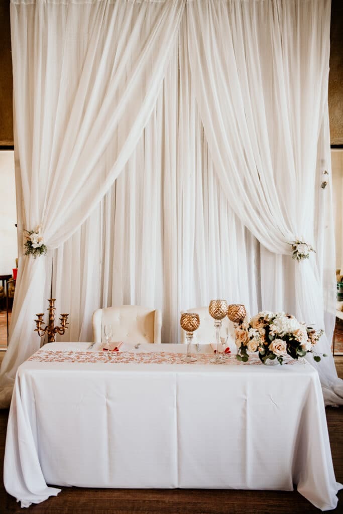 sweetheart table draped in white with gold accents and tall white drapes behind it by Making It Matthews