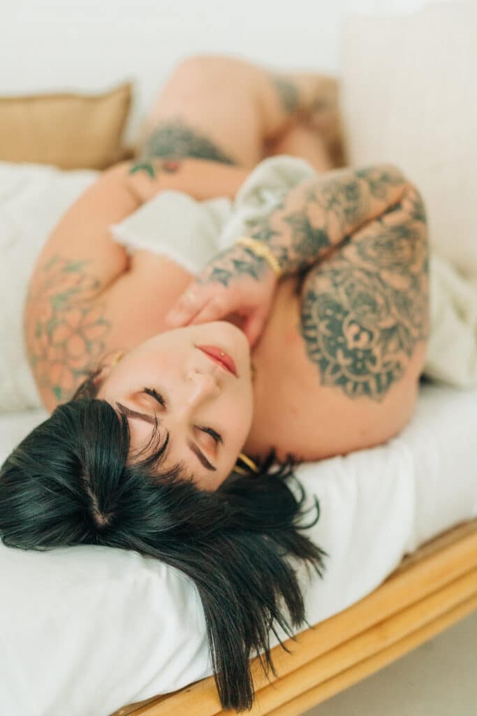 woman with black hair and tattoos lying on white sheets by Sydney Morman Boudoir Photography