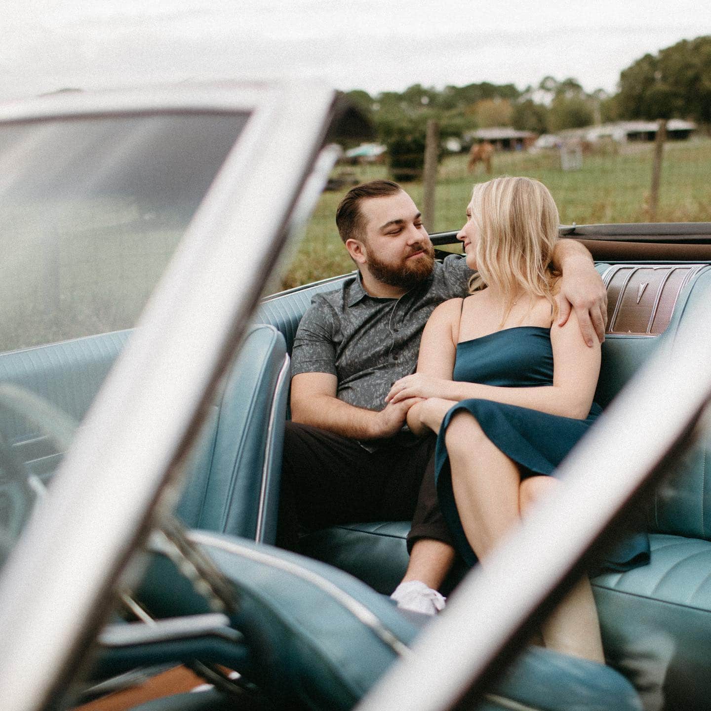 blonde girl wearing emerald dress sits next to man with beard in the backseat of classic car