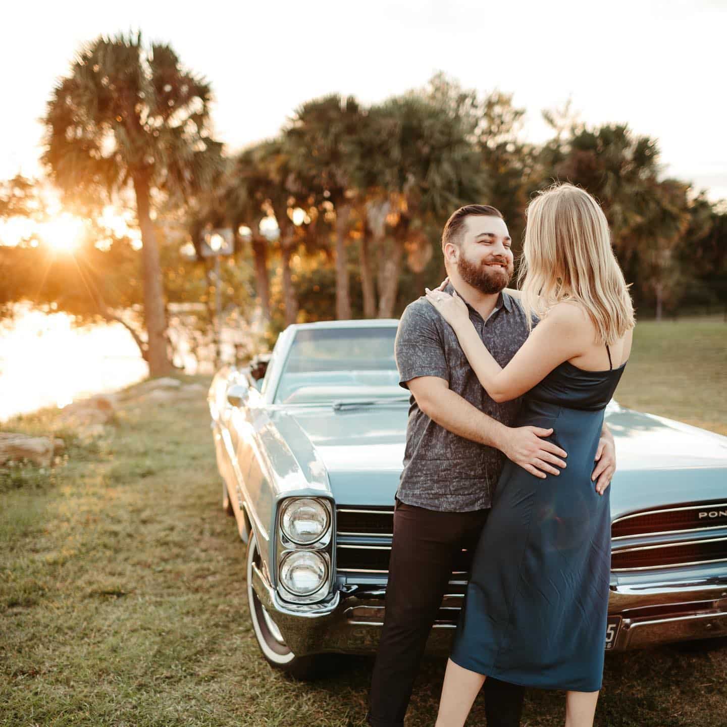 man holds woman by hips and poses for picture in front of classic convertible car near lake at sunset