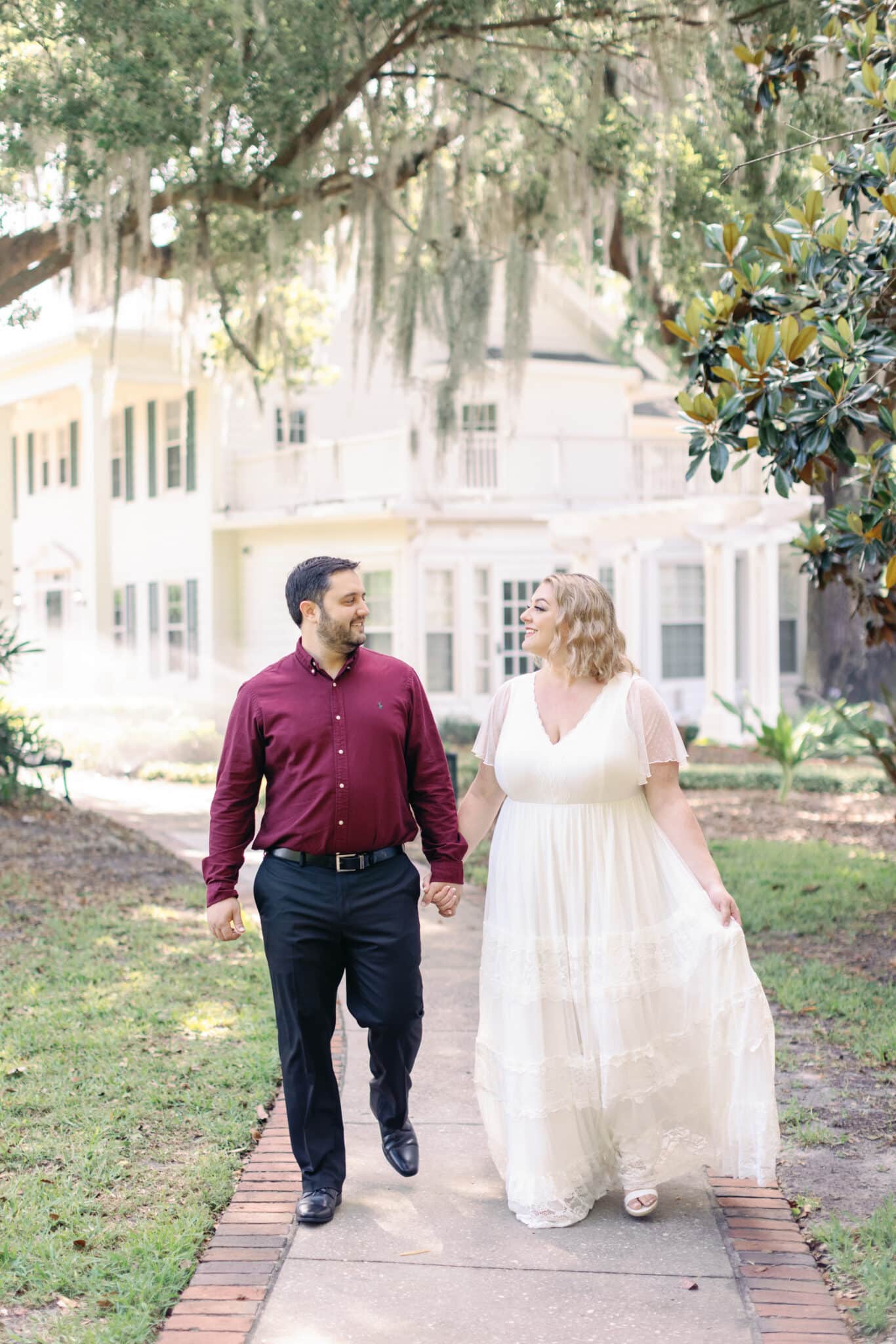 woman in long white dress walk on sideway holding hands and looking at man wearing red long sleeve shirt with black pants under cypress tree