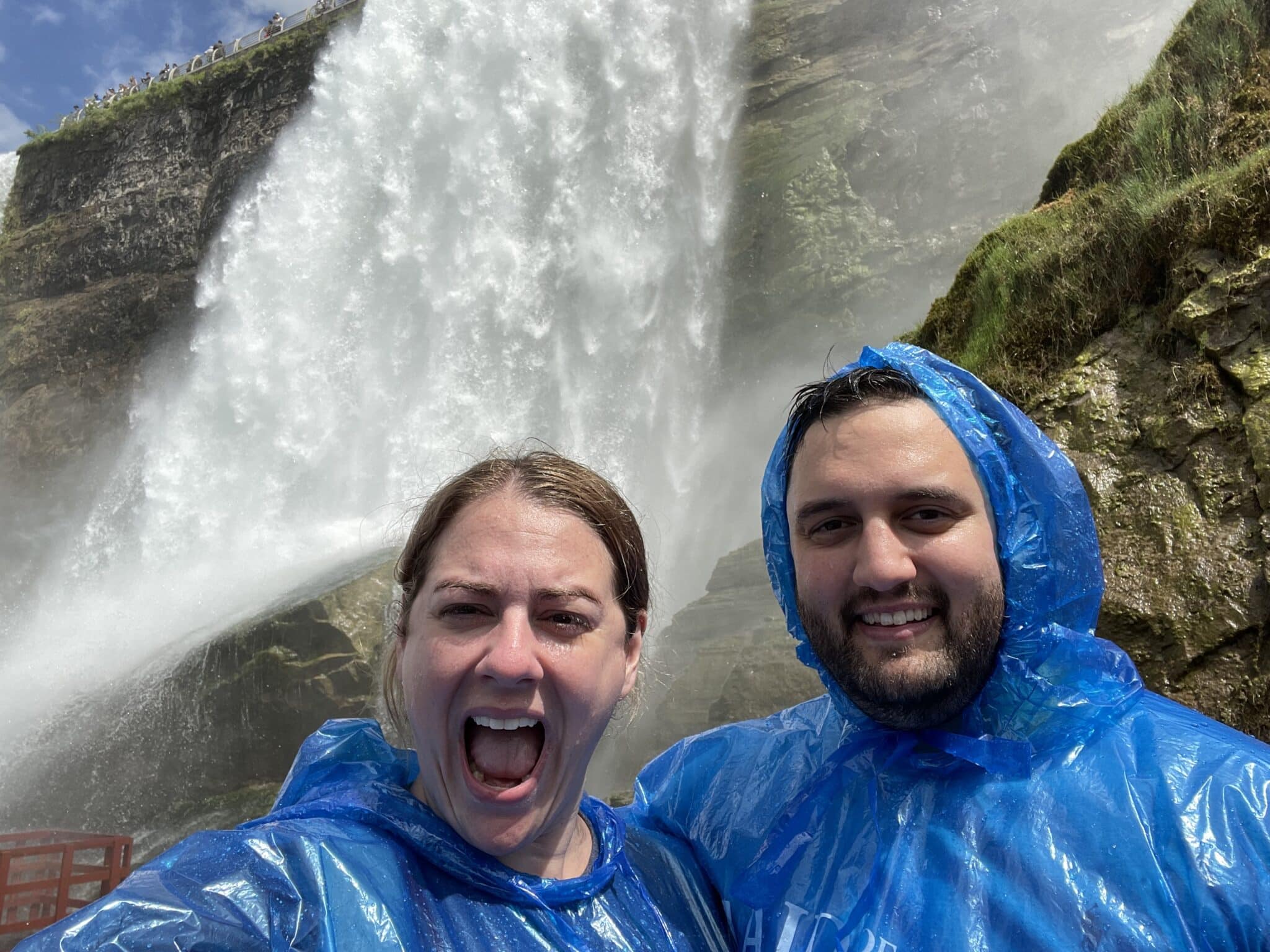 man and woman looking at camera man smiling and woman with mouth open in surprise with big waterfall behind them as they wear blue ponchos