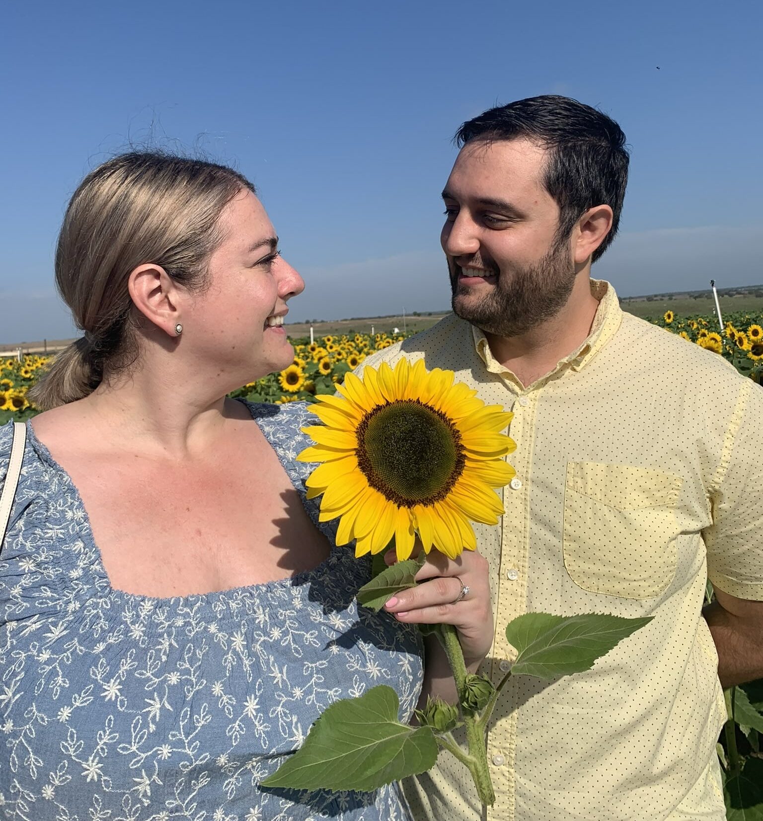 woman wearing blue top holds big sunflower as she looks to the right at man looking back smiles at her wearing yellow shirt with sunflower field in background