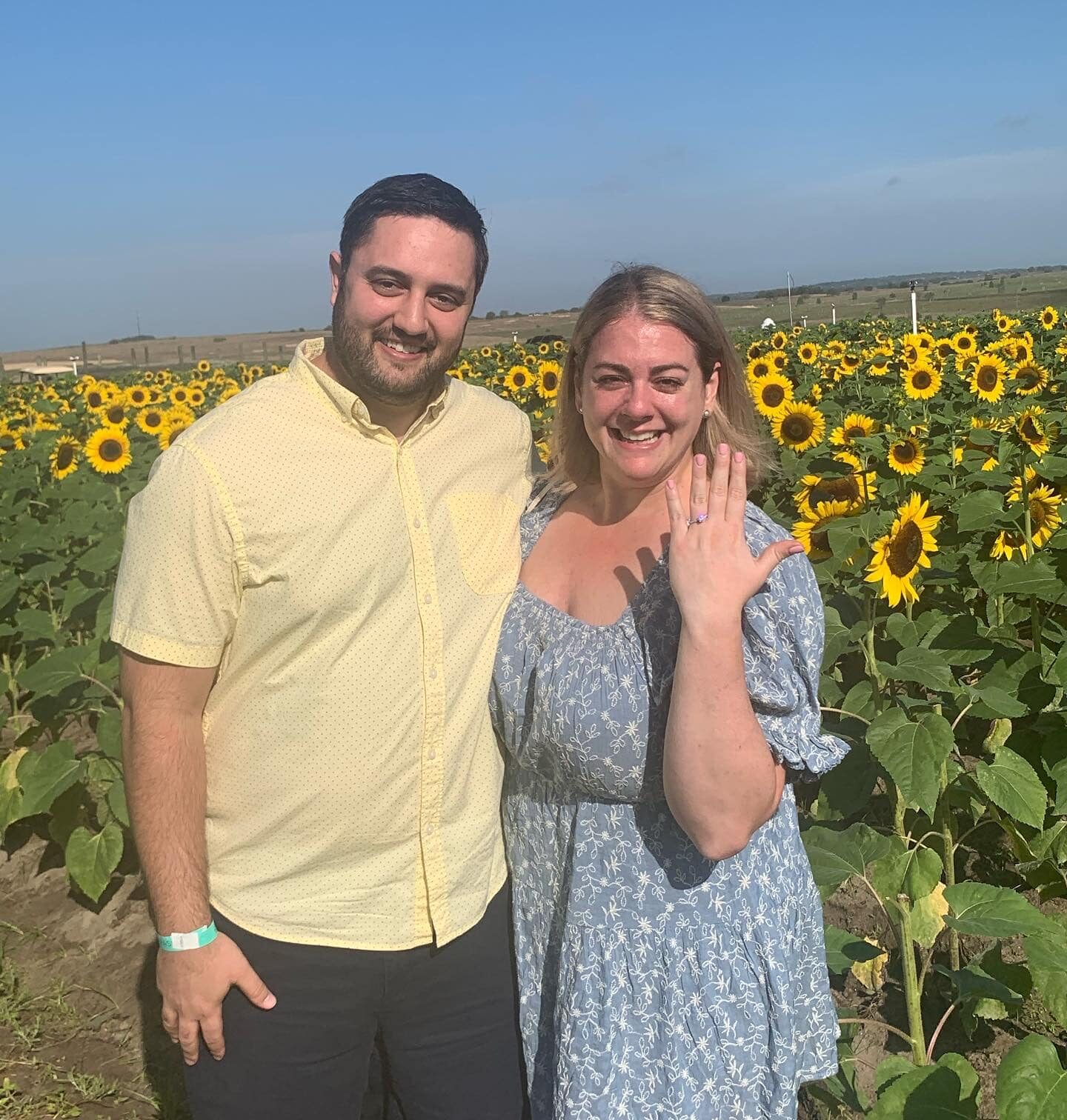 couple stand together in front of southern hills sunflower farm while woman holds hand up with engagement right and smiles