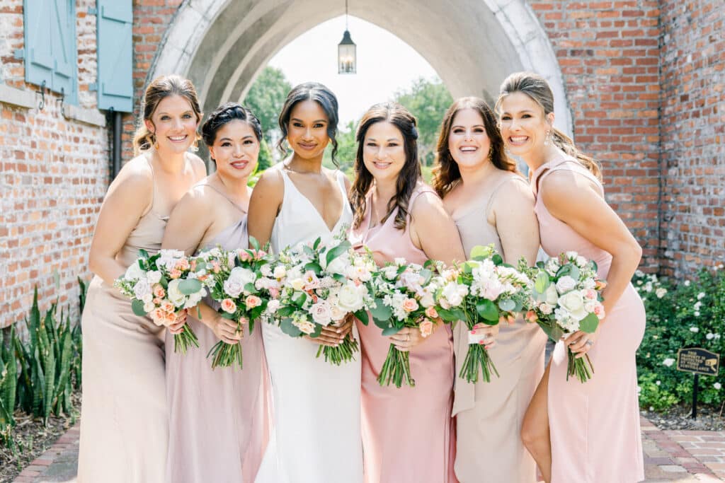 bride with bridesmaids in ombre shades of pink and taupe under stone archway