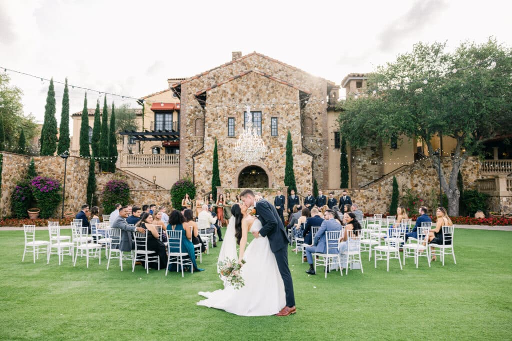 bride and groom kissing after ceremony at the end of an outdoor aisle against a stone backdrop building