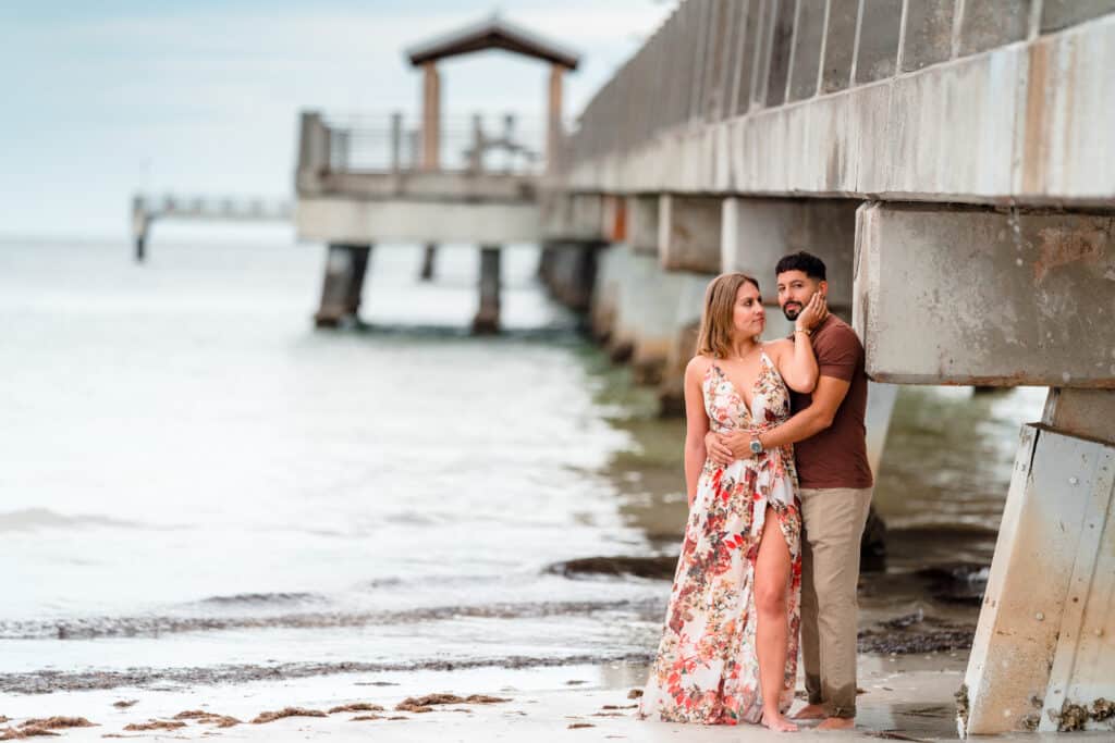 couple posing for a photo on the beach below a pier photo by Jerzy Nieves Photography LLC