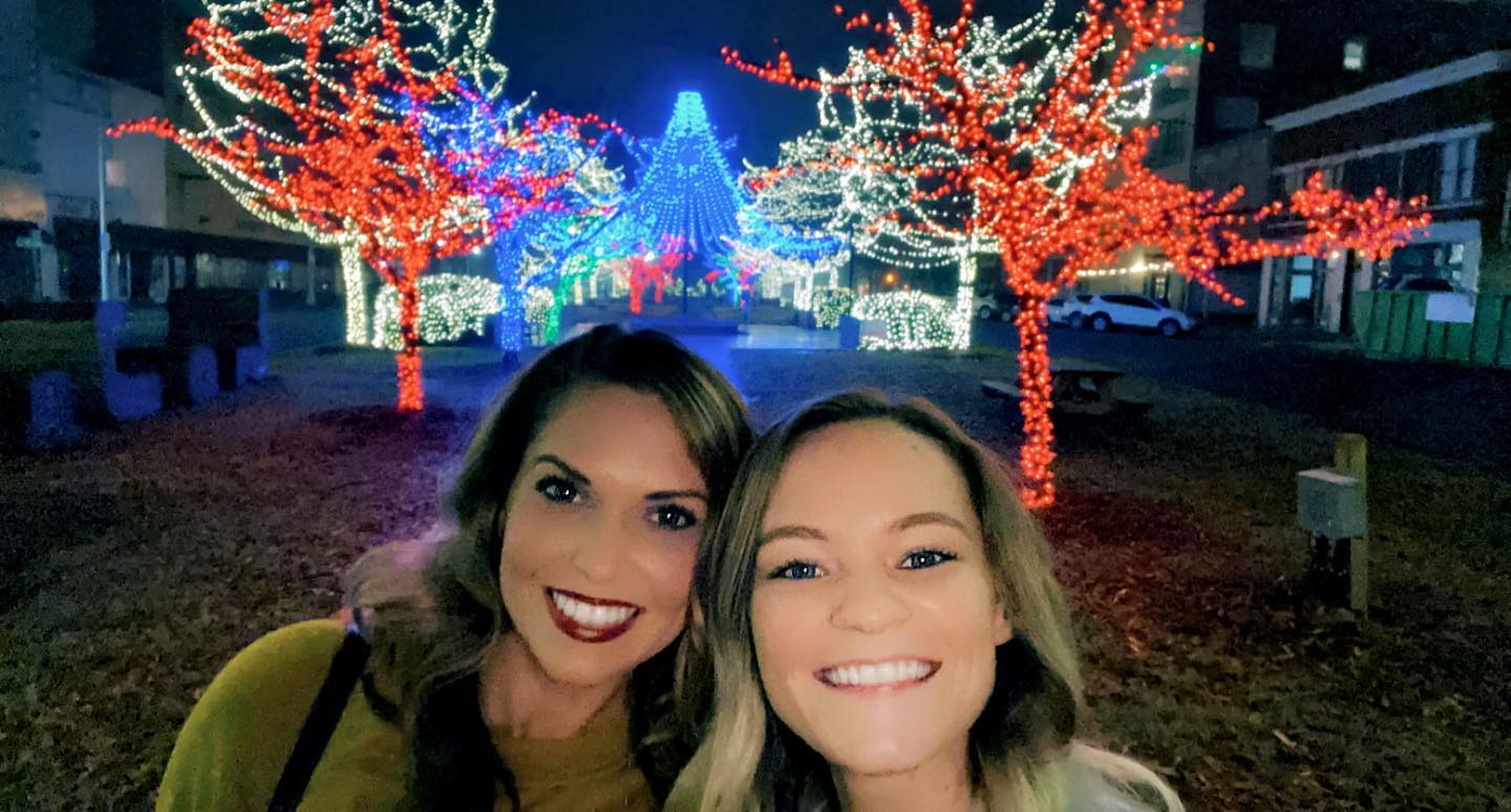 couple stands together for picture in front of trees with christmas light decorating them