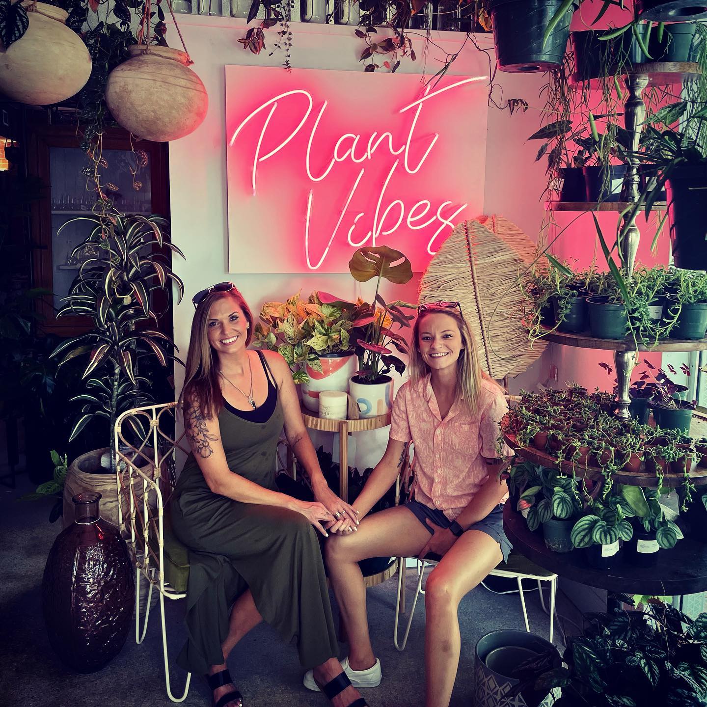 two girls sit in chairs surrounded by plants in front of a pink neon sign that reads plant vibes