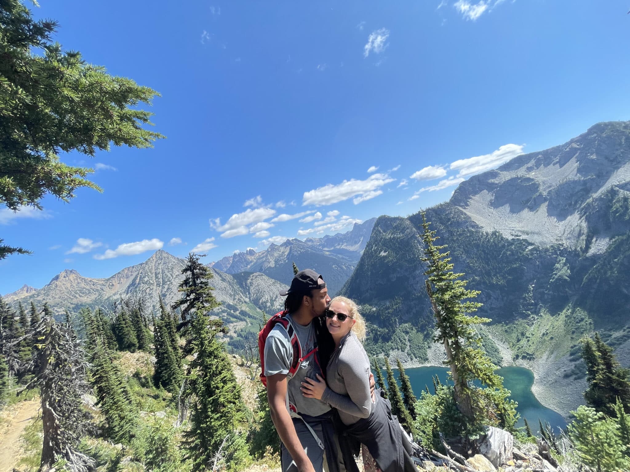 couple stands together while on a hike at the top of mountain overlooking other mountain ridge and lake