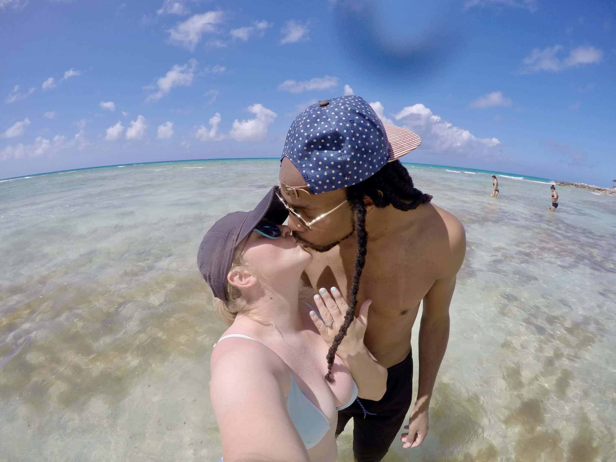 man and woman kiss while wearing hats standing in the ocean on a beach while girl holds up hand to show engagement ring from her Carnival Cruise Marriage Proposal