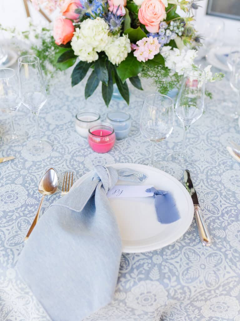 place setting with lace linen and white plate with silverware on each side and light blue napkin tied in a knot with floral arrangement above it