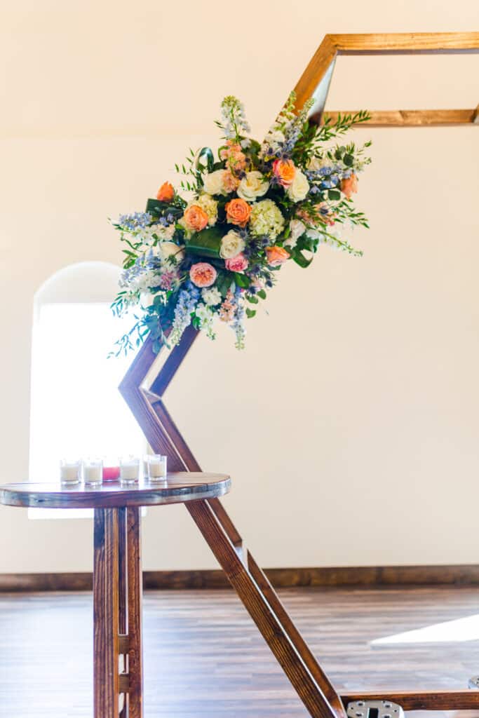 close up image of floral arrangement on wooden hexagon arch with wooden table to the left of it holding votive candles