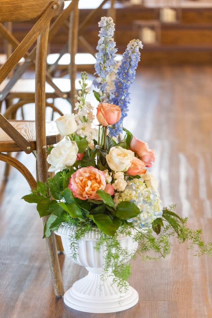 close up of floral arrangement sitting on wooden floor next to wooden chair inside of a white base