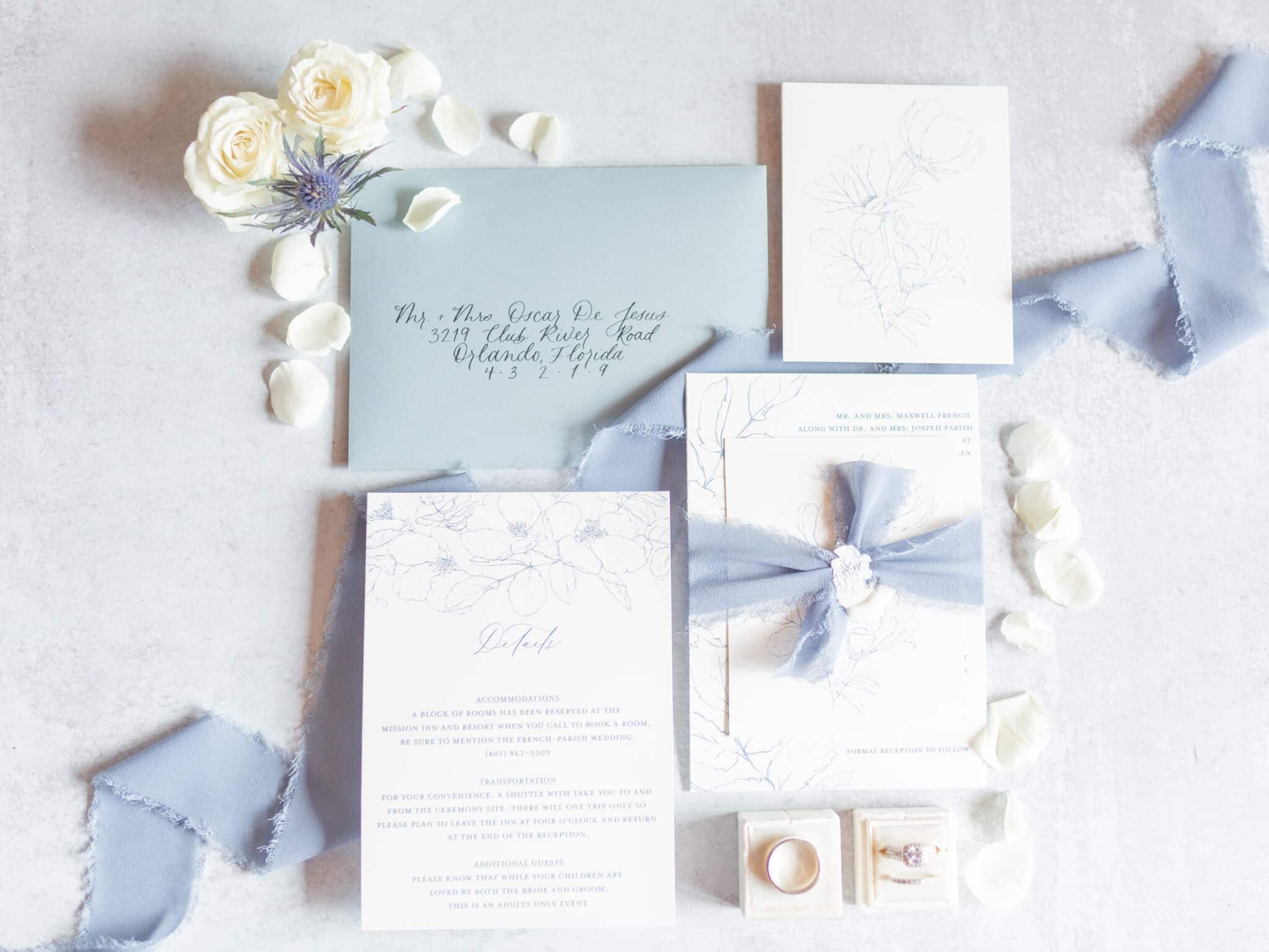 event stationary with invitation envelop and other items with hand calligraphy and dusty blue ribbon with ring boxes and flower petals