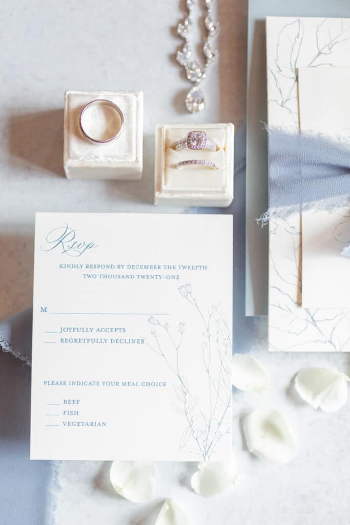wedding bands and engagement ring inside of velvet boxes next to rsvp card for wedding
